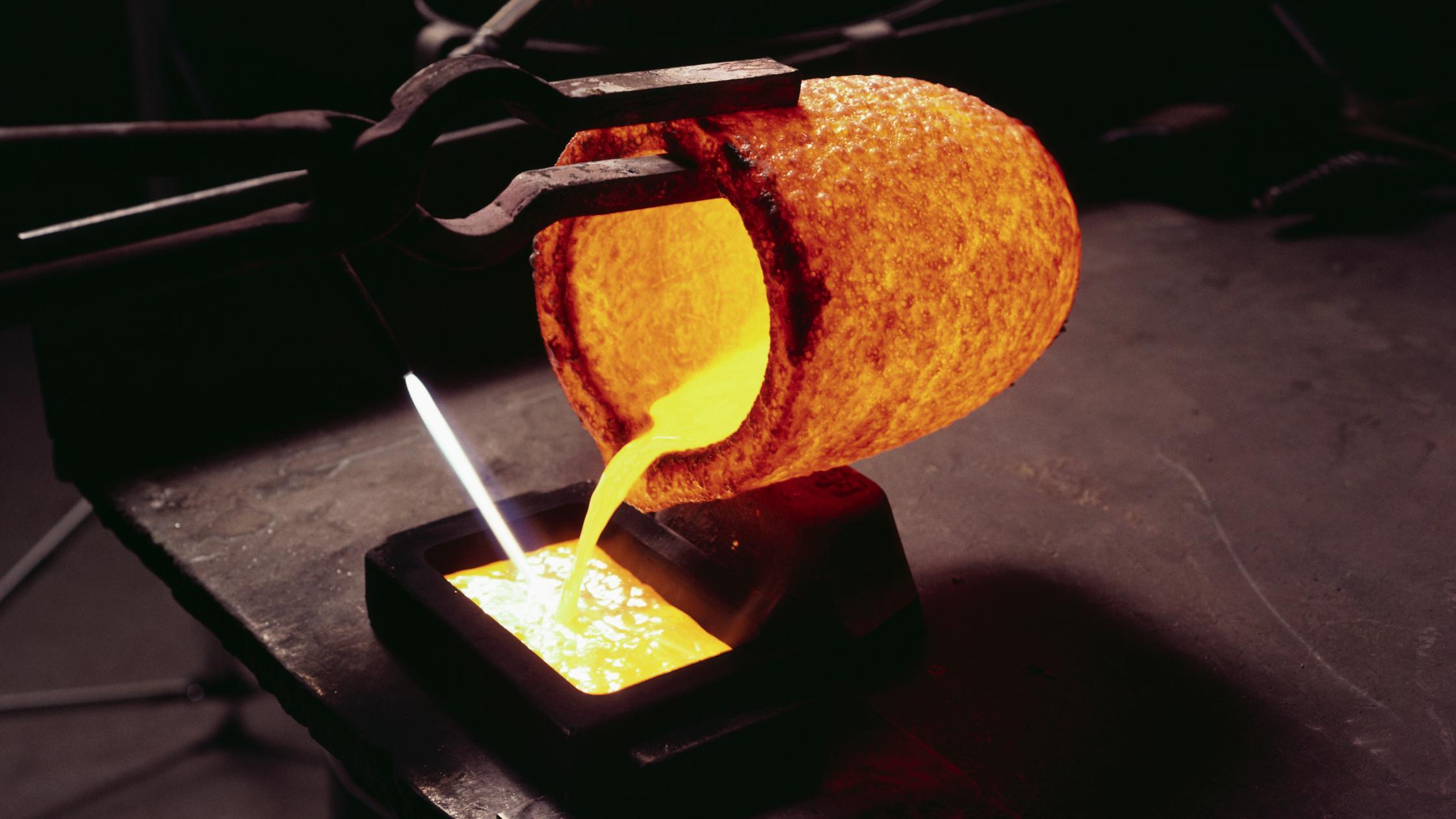 Molten gold pours into a bar mold from a red-hot crucible held by tongs.